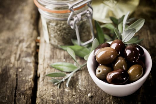 A bowl of fresh black olives and container of dried herbs stand on an old wooden kitchen table for use as ingredients in cooking