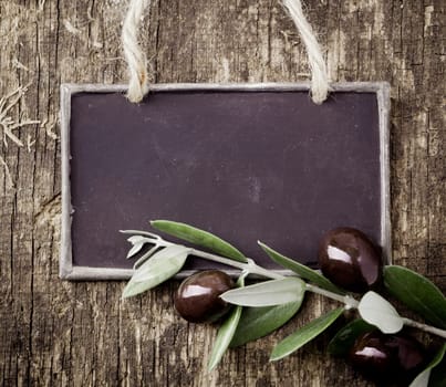Fresh black olives on a leafy twig and a blank slate for your text lying on an old weathered wooden table