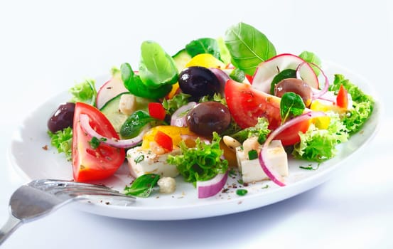 Healthy mixed Greek salad served on a white plate with silver fork containing crisp leafy greens, olives, feta, onion , tomato, cucumber and radish