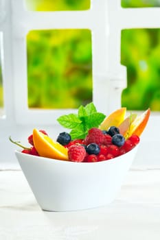 Bowl of fresh berries including raspberries, blueberries and redcurrants, garnished with sliced oranges and mint on an indoor table with copyspace