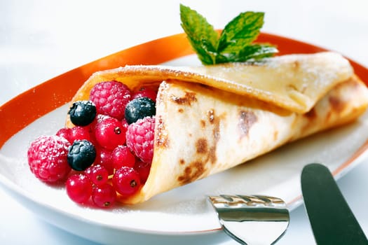 Golden pancake filled with raspberries, redcurrants and blueberries served with a knife and fork for a scrumptious dessert