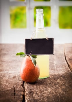 A juicy ripe red pear stands alongside a bottle of natural freshly squeezed pear juice with a blank slate on display on a wooden table