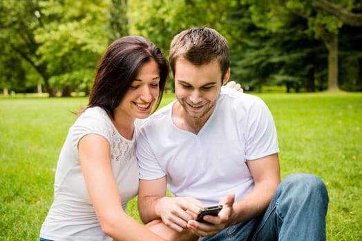 Young happy couple sitting on grass and and looking on smartphone display