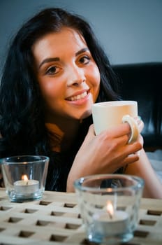 Head and shoulders shot of a young black-haired woman sitting behind a table and holding a coffee cup with candles in the foreground,looking at the camera