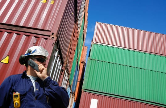 stacks of cargo and shipping containers with dock worker talking in phone