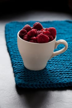 Closeup of fresh raspberries in white cup on a blue mat with shaddow background