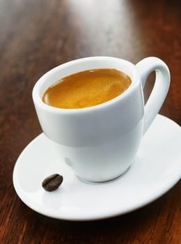 A full white coffee cup with a single coffee bean on the saucer, on a dark wood background