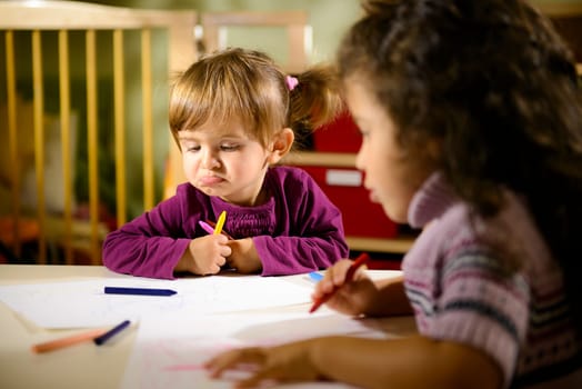Children having fun at school, two young girls drawing in kindergarten with sad child contemplating her drawing