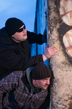 two middle-aged men look crouched around a concrete wall
