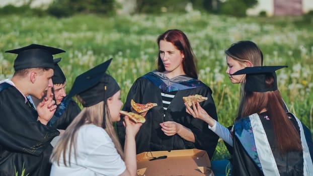 Graduates in black suits eating pizza in a city meadow