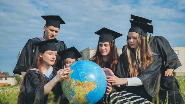 Graduates in black robes examine a geographical globe sitting on the grass