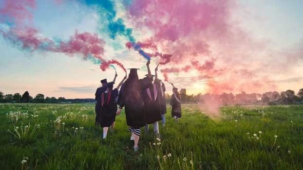 Graduates in costume walk across a field with coloured red and blue smoke at sunset