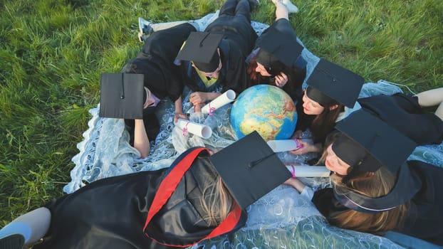 Graduates in black robes looking at a georgraphic globe lying on the grass