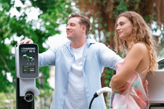 Happy and lovely couple with eco-friendly conscious recharging electric vehicle from EV charging station. EV car technology utilized as alternative transportation for future sustainability. Synchronos