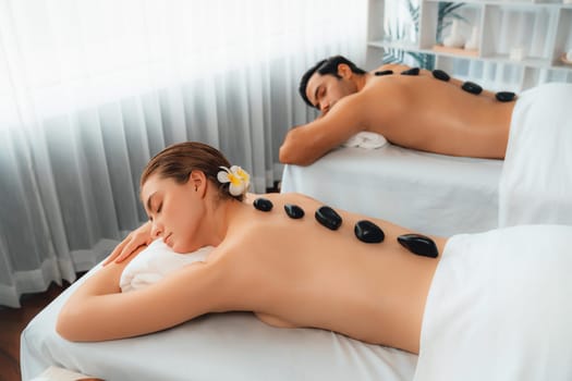 Hot stone massage at spa salon in luxury resort with day light serenity ambient, blissful couple customer enjoying spa basalt stone massage glide over body with soothing warmth. Quiescent