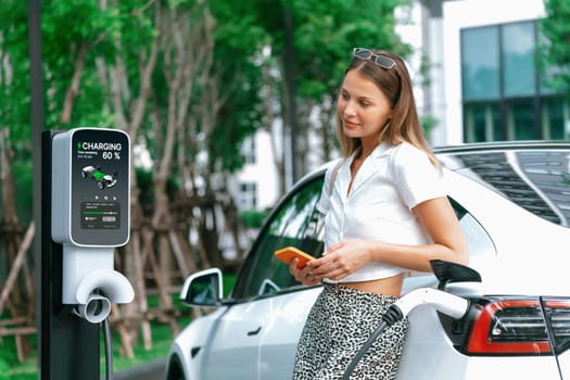 Young woman travel with EV electric car charging in green sustainable city outdoor garden in summer. Urban sustainability lifestyle by green clean rechargeable energy of electric BEV vehicle innards