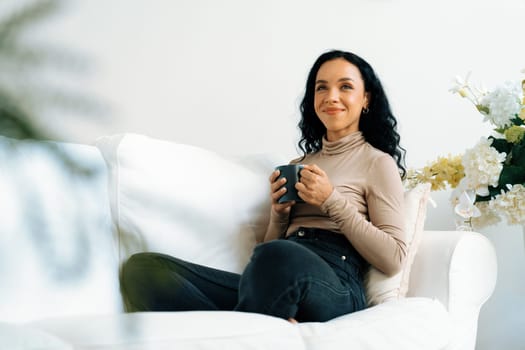 Happy woman drinking coffee on a sofa at home for crucial rest and relaxation. Portrait of young African American woman holding a cup.