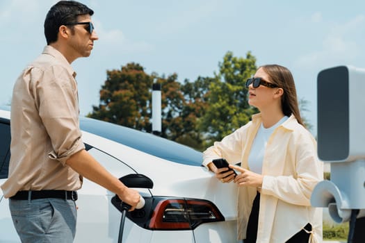 Young couple use smartphone to pay for electricity at public EV car charging station green city park. Modern environmental and sustainable urban lifestyle with EV vehicle. Expedient