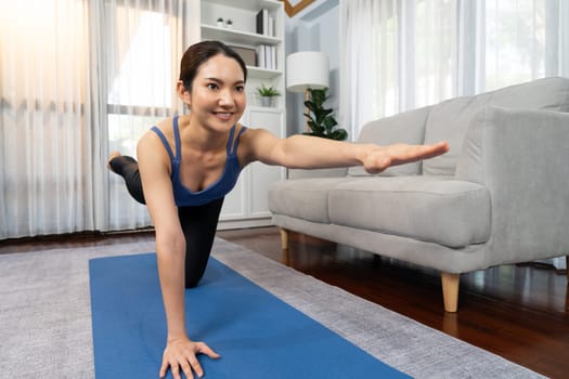 Asian woman in sportswear doing yoga exercise on fitness mat as her home workout training routine. Healthy body care and calm meditation in yoga lifestyle with comfortable and relaxation. Vigorous