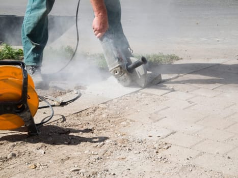 Legs of a worker with cut-off grinder and cable drum cutting pavement slabs in daylight and rising sanding dust