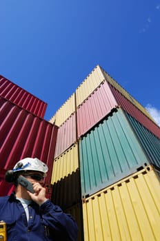 dock worker talking in phone, large stacks of shipping containers in background