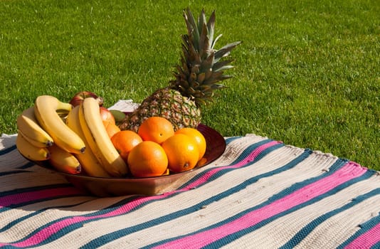 fruit bowl with apples, bananas, oranges and pineapple on a colorful picnic blanket (grass field background)