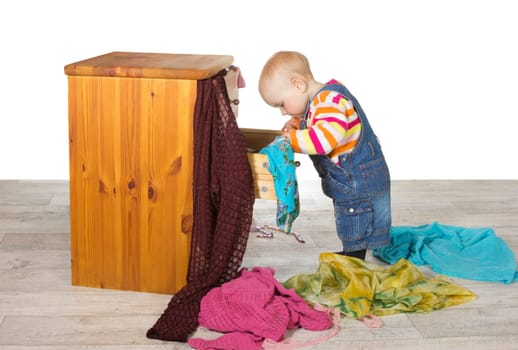 Happy baby standing searching in a drawer of a small wooden chest of drawers unpacking colourful clothes and textiles onto the floor in a random pile