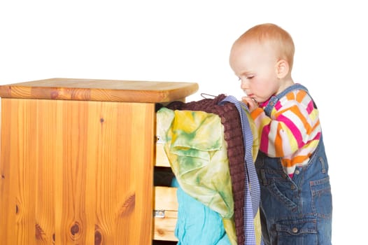 Curious little baby standing peering into a drawer with colourful textiles spilling out onto the floor on a white background