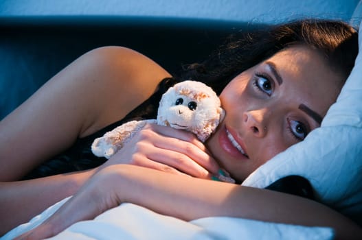 Head and shoulders shot of a young black-haired woman lying sideways on a sofa with small cuddly toy in her arms and looking at the camera
