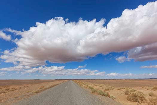 Road with desert landscape and cloudy blue sky in the Sahara desert of Morocco.