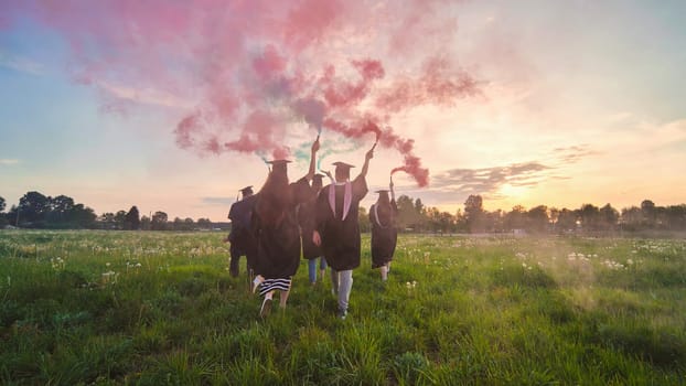 Graduates in costume walk with a smoky multi-colored smoke at sunset.