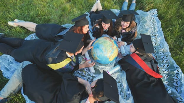 Graduates in black robes looking at a georgraphic globe lying on the grass
