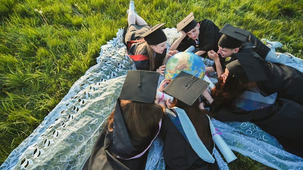 Graduate students in black robes study a globe on the grass