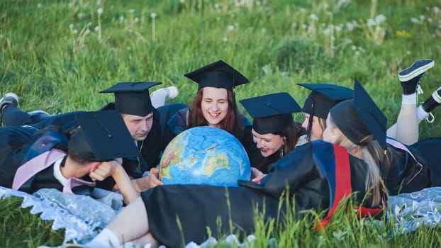 Graduates in black robes looking at a georgraphic globe lying on the grass