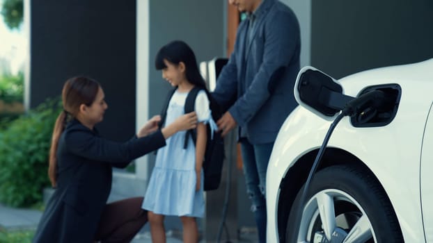 Progressive young parents and daughter with electric vehicle and home charging station. Green and clean energy from electric vehicles for healthy environment. Eco power from renewable source.