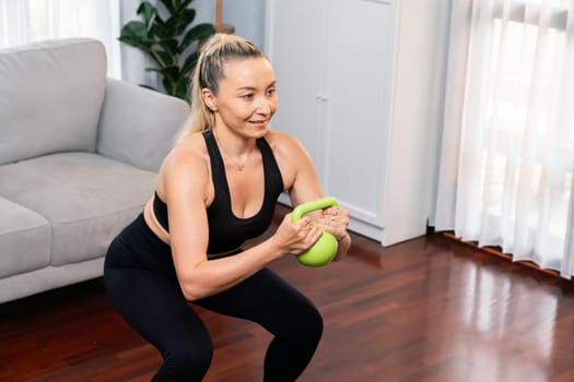 Athletic and sporty senior woman engaging in leg day training session with squat and bodyweight kettle ball at home exercise as concept of healthy fit body lifestyle after retirement. Clout