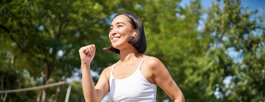 Smiling fitness girl, asian runner in wireless headphones, running in park, jogging workout outdoors. Sport and wellness concept