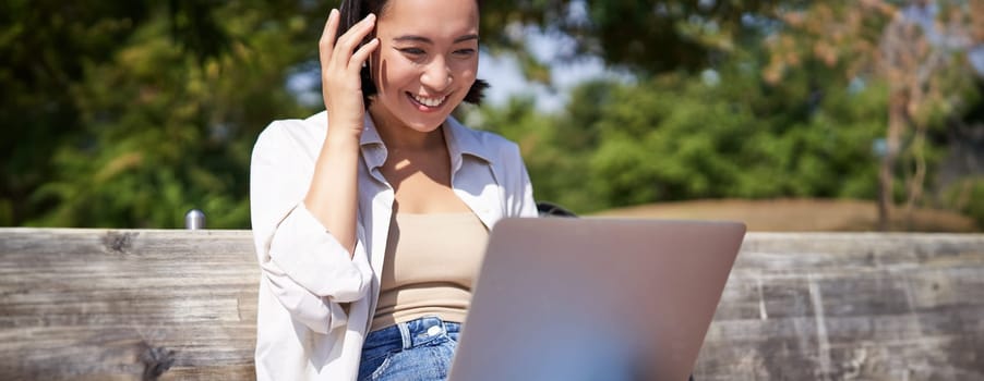 Beautiful asian girl laughing, watching video on laptop, listening music or video chatting online while sitting in park on bench.