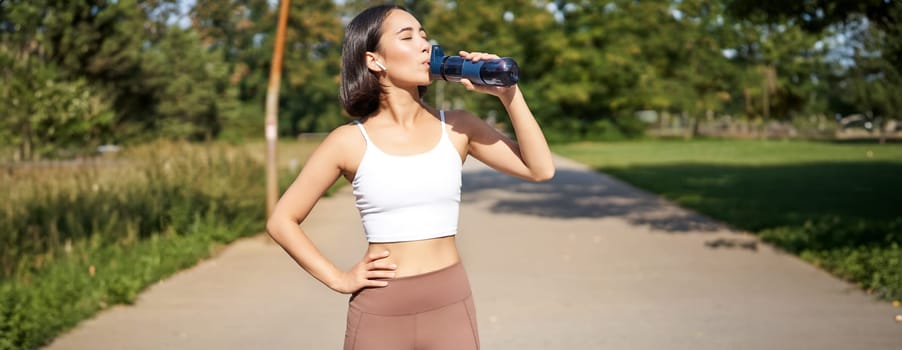 Happy asian sportswoman, runner drinks water from bottle while running, workout on fresh air in park.