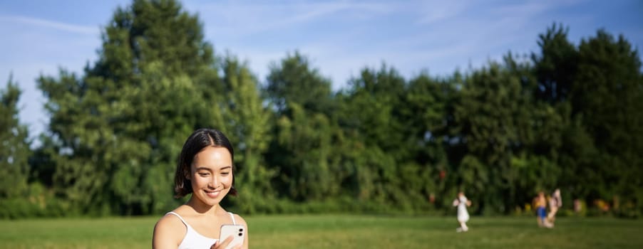 Portrait of asian girl sitting on mat in park, talking to smartphone, meditating online with yoga instructor on fresh air.