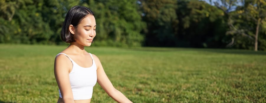 Woman meditating in park on rubber mat, sitting on green lawn and practice yoga, concept of sport and wellbeing.