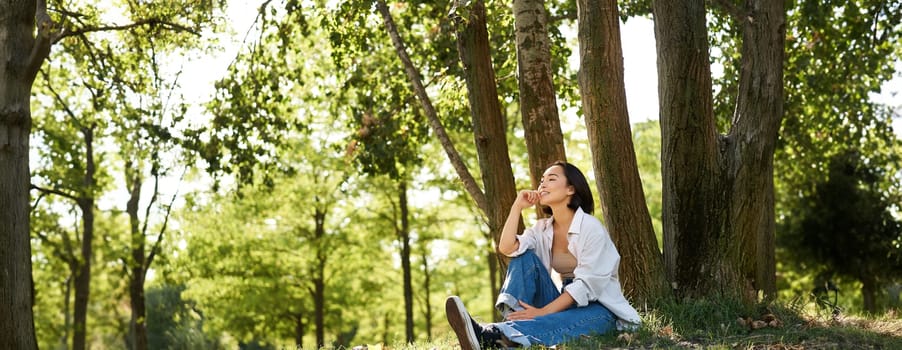 Relaxed young woman, resting near tree, sitting in park on lawn under shade, smiling and looking happy, walking outdoors on fresh air.