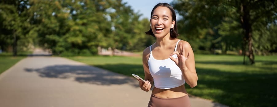 Sport people. Happy asian girl, sportswoman showing okay sign and smiling, workout in gym, say yes, approve workout outdoors.