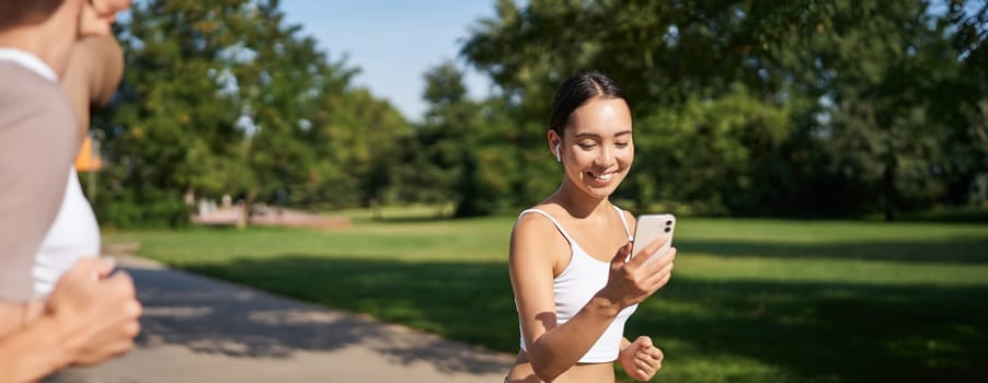 Happy asian fitness girl running and checking her stats, daily goals on smartphone app. Young woman jogging and looking at mobile screen.