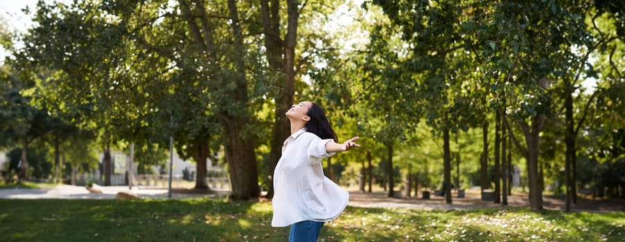 Carefree asian girl dancing, feeling happiness and joy, enjoying the sun on summer day, walking in park with green trees.