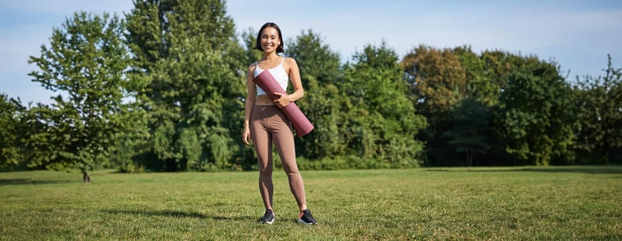 Confident and sporty young asian woman, standing in park with rubber mat, wearing sportswoman, smiling at camera, workout on fresh air.