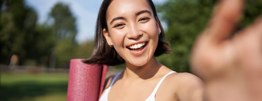 Happy asian fitness girl takes selfie with rubber yoga mat in park. Healthy young sportswoman makes photo of herself during workout.