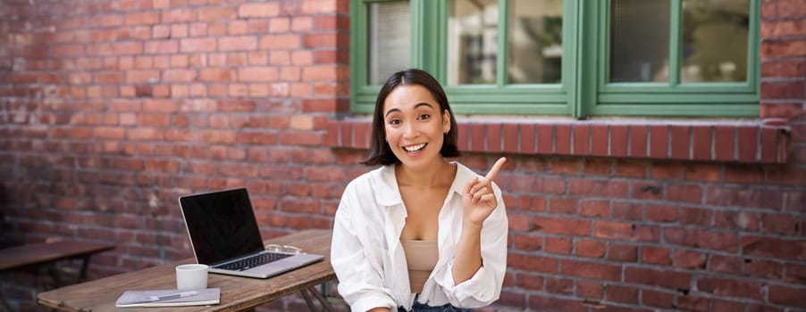 Asian girl with surprised face, sitting with laptop, pointing left at copy space, banner with information, looking amazed.