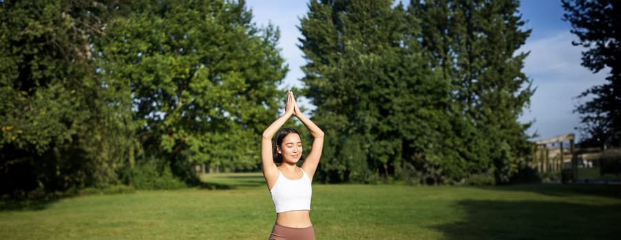 Vertical shot of asian woman standing in asana, doing yoga exercises on fresh air in park, wearing leggings, standing on rubber mat.