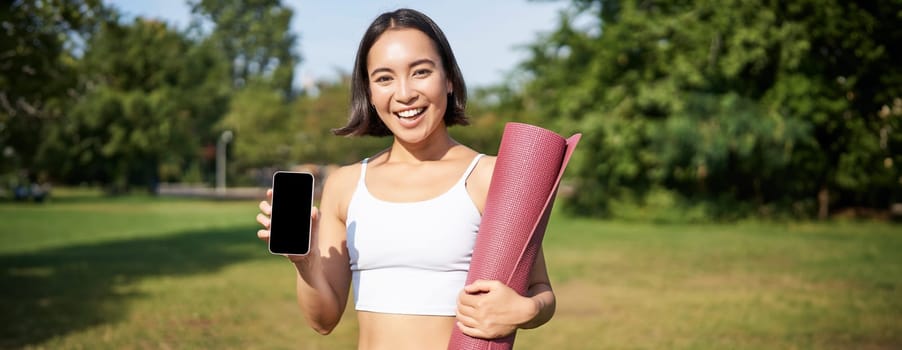 Smiling asian fitness girl with rubber yoga mat, shows her smartphone screen, recommends workout application, stands on lawn in park.